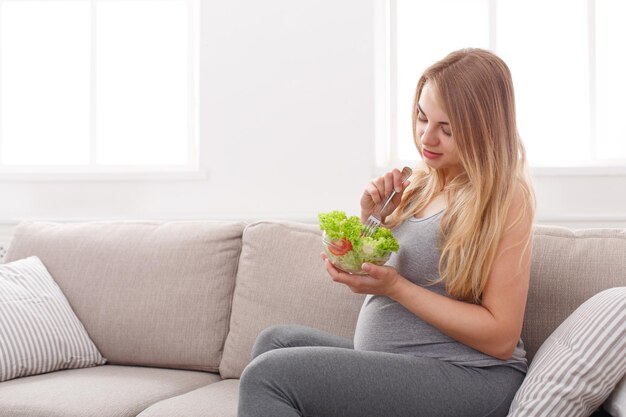 Jeune femme enceinte mangeant de la salade verte. Jolie blonde enceinte assise sur un canapé et ayant une collation fraîche. Concept de nutrition saine et de grossesse