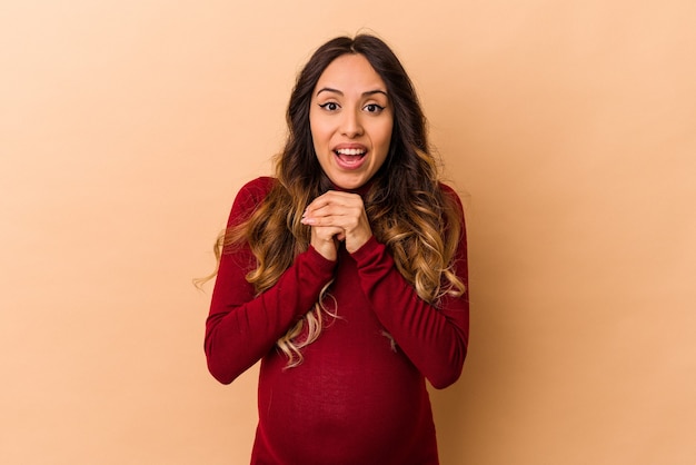 Jeune femme enceinte mexicaine isolée sur fond beige priant pour la chance, étonné et ouvrant la bouche à l'avant.