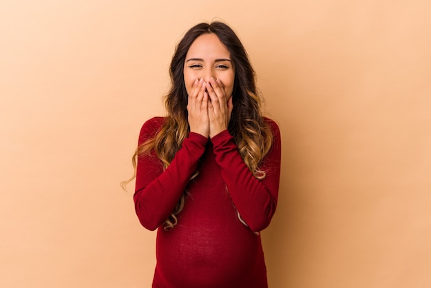 Jeune femme enceinte mexicaine isolée sur fond beige en riant de quelque chose, couvrant la bouche avec les mains.