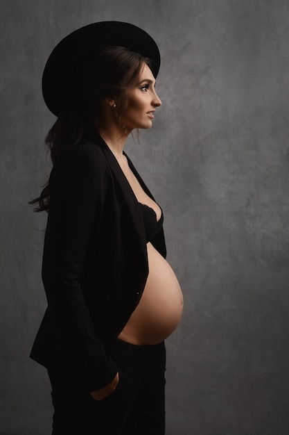 Jeune femme enceinte à la mode dans un chapeau à la mode et une tenue noire à la mode avec un ventre nu...