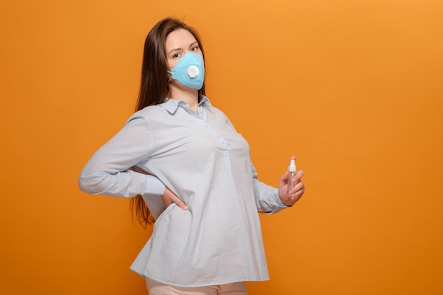 Jeune femme enceinte sur un mur orange dans un masque médical de protection et un antiseptique en spray à la main