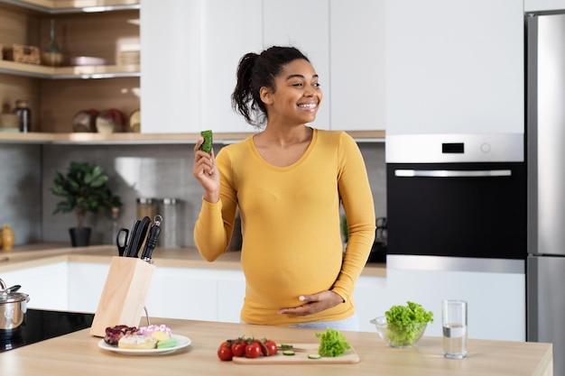 Une jeune femme enceinte noire satisfaite avec un gros ventre mange des aliments de cuisson de légumes biologiques à table avec de la salade et des bonbons