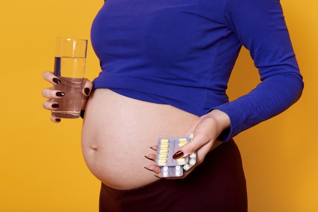 Jeune femme enceinte sans visage tenant un verre d'eau et de pilules, debout isolé sur jaune