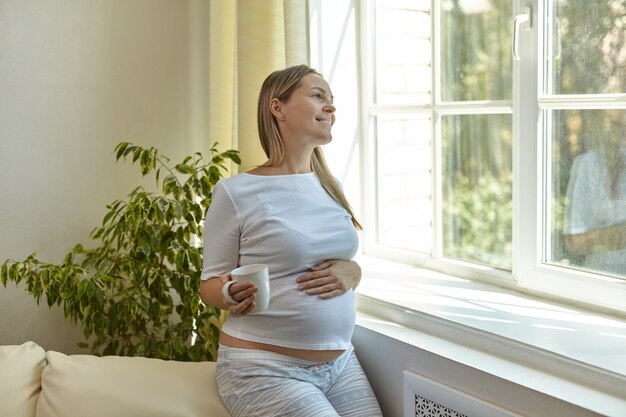 Une jeune femme enceinte se tient près de la fenêtre à la maison avec une tasse de café