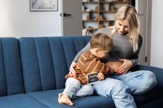 Une jeune femme enceinte et son fils sont assis sur un canapé à la maison et un garçon tient une échographie à la main