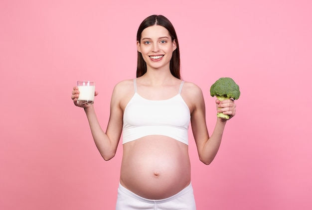 Jeune femme enceinte souriante et joyeuse, femme aux cheveux bruns, tient un verre de lait et de brocoli à la main. Une femme enceinte suit un régime, prend soin de la santé de son bébé à naître.