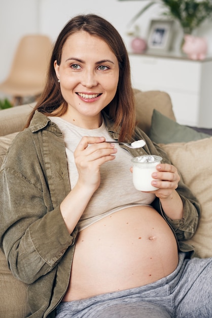 Jeune femme enceinte souriante en tenue décontractée, manger du yaourt tout en vous relaxant sur le canapé et profiter du temps libre à la maison