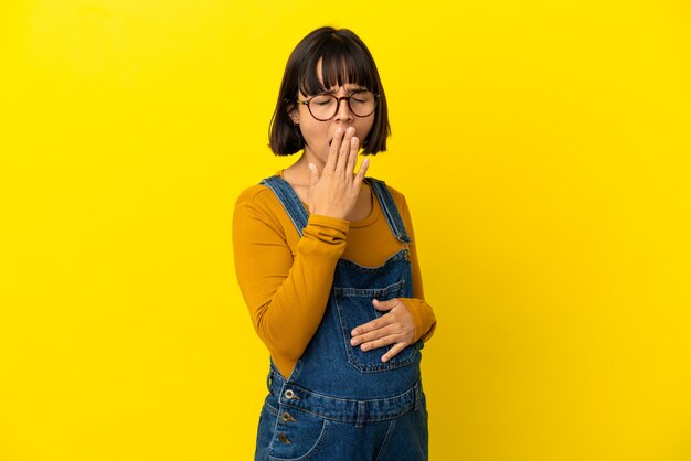 Jeune femme enceinte sur une surface jaune isolée bâillant et couvrant la bouche grande ouverte avec la main