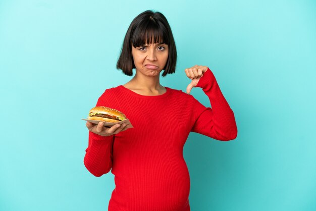 Jeune femme enceinte tenant un hamburger sur fond isolé montrant le pouce vers le bas avec une expression négative