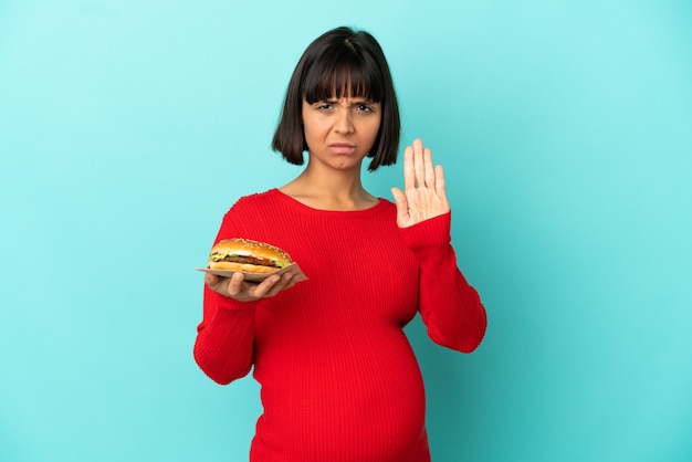 Jeune femme enceinte tenant un hamburger sur un mur isolé faisant un geste d'arrêt