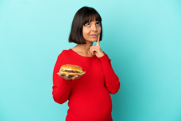 Photo jeune femme enceinte tenant un hamburger sur une surface isolée ayant des doutes en levant les yeux