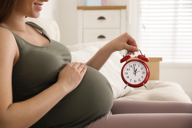 Jeune femme enceinte tenant un réveil près de son ventre à la maison gros plan Temps de donner naissance