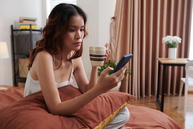 Photo jeune femme endormie assise dans son lit, buvant du café et faisant défiler les médias sociaux