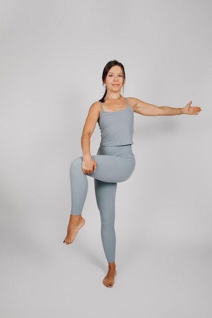 Jeune femme engagée dans le sport Brunette en costume de fitness effectue des exercices de yoga ou d'étirement sur fond gris isolé, elle se tient sur une jambe, sa main levée à côté concept de mode de vie sain