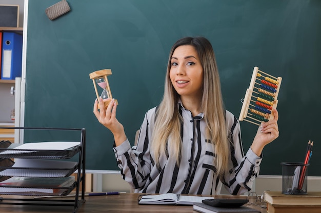 jeune femme enseignante assise au bureau de l'école devant le tableau noir dans la salle de classe expliquant la leçon tenant un boulier et un sablier souriant heureux et heureux