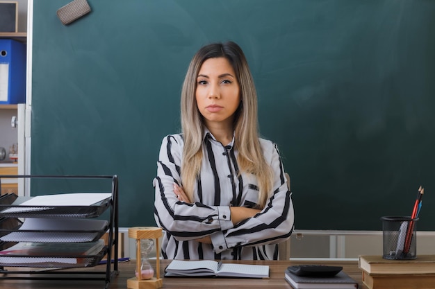 jeune femme enseignante assise au bureau de l'école devant le tableau noir dans la salle de classe vérifiant les devoirs des élèves croisant les mains sur sa poitrine avec un visage sérieux regardant la caméra