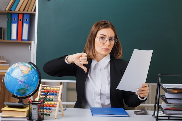 Jeune femme enseignante portant des lunettes assis au bureau de l'école avec globe et livres devant le tableau noir dans la salle de classe tenant une feuille de papier vide blanche montrant le pouce vers le bas à l'air mécontent