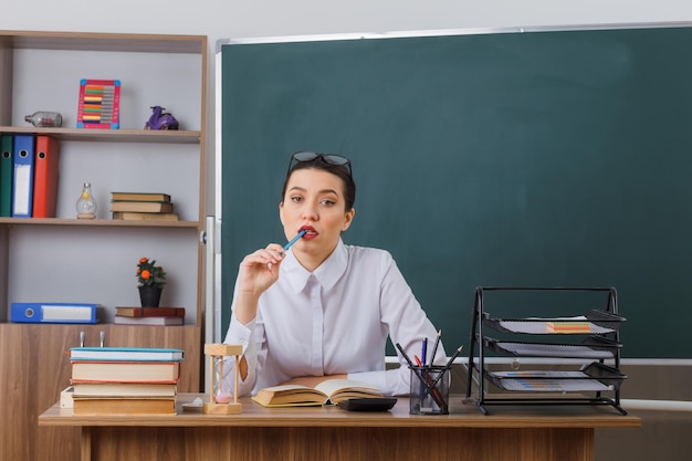 Jeune femme enseignante portant des lunettes assis au bureau de l'école avec un livre devant le tableau noir dans la salle de classe à la pensée perplexe