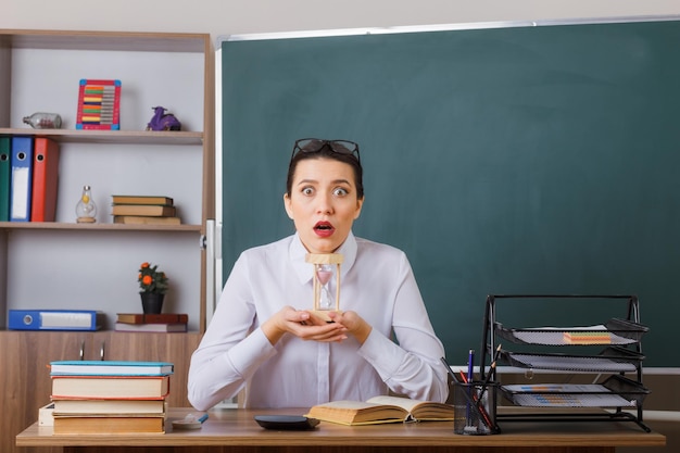 Jeune femme enseignante portant des lunettes assis au bureau de l'école avec un livre devant le tableau noir dans la salle de classe présentant un sablier étonné et surpris