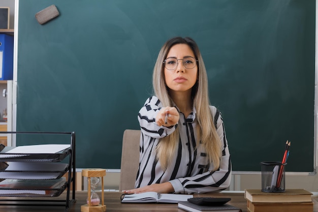 jeune femme enseignante portant des lunettes assise au bureau de l'école devant le tableau noir dans la salle de classe expliquant la leçon pointant avec l'index vers la caméra avec un visage sérieux