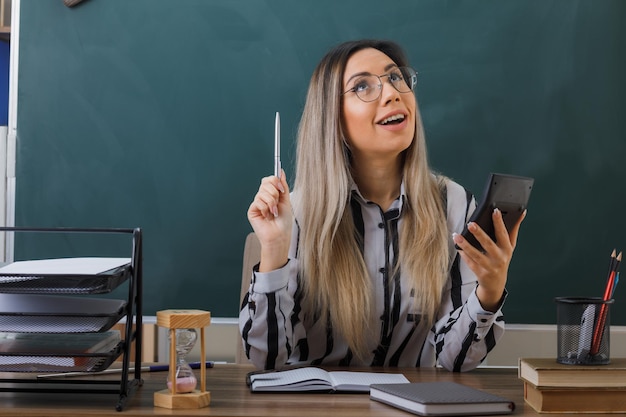 jeune femme enseignante portant des lunettes assise au bureau de l'école devant le tableau noir dans la salle de classe expliquant la leçon tenant la calculatrice levant les yeux avec une expression pensive pensant