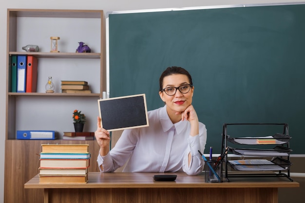 Jeune femme enseignante portant des lunettes montrant un petit tableau regardant la caméra souriant confiant assis au bureau de l'école devant le tableau noir dans la salle de classe