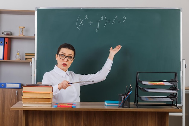 Jeune femme enseignante portant des lunettes tenant un pointeur tout en expliquant la leçon à l'air confiant assis au bureau de l'école devant le tableau noir dans la salle de classe