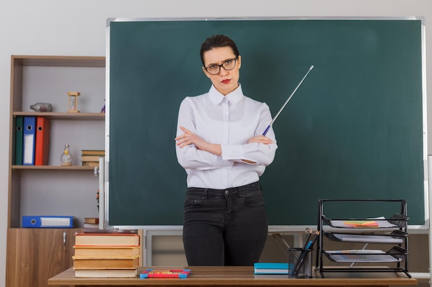 Jeune femme enseignante portant des lunettes tenant un pointeur tout en expliquant la leçon croisant les mains sur la poitrine debout au bureau de l'école devant le tableau noir dans la salle de classe