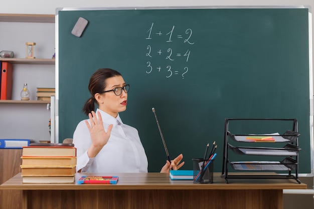 Jeune femme enseignante portant des lunettes tenant un pointeur tout en expliquant la leçon semblant confuse faisant un geste d'arrêt avec la main assise au bureau de l'école devant le tableau noir dans la salle de classe