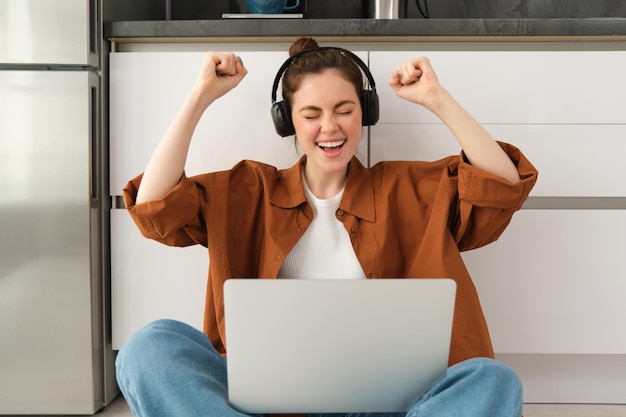 Jeune femme enthousiaste atteint l'objectif s'assoit sur le sol avec un ordinateur portable et des écouteurs crie avec