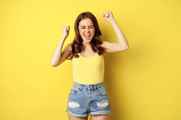 Jeune femme enthousiaste criant et applaudissant, se réjouissant, levant les mains et criant de joie, chantant, debout sur fond jaune