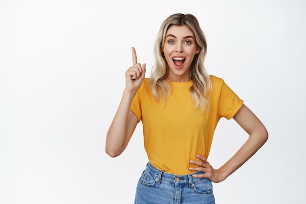 Jeune femme enthousiaste pointant le doigt vers le haut et souriante, intriguée par la publicité recommandant un magasin ou une promotion de vente fond blanc