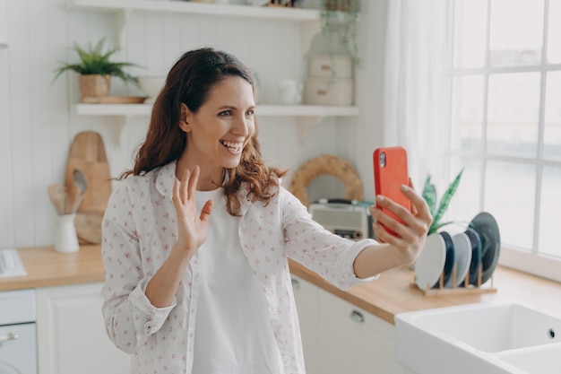 Jeune femme espagnole a un appel vidéo sur le téléphone à la cuisine La fille agite la main et sourit