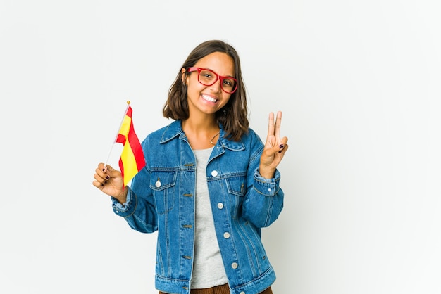 Jeune femme espagnole tenant un drapeau isolé sur blanc montrant le signe de la victoire et souriant largement.