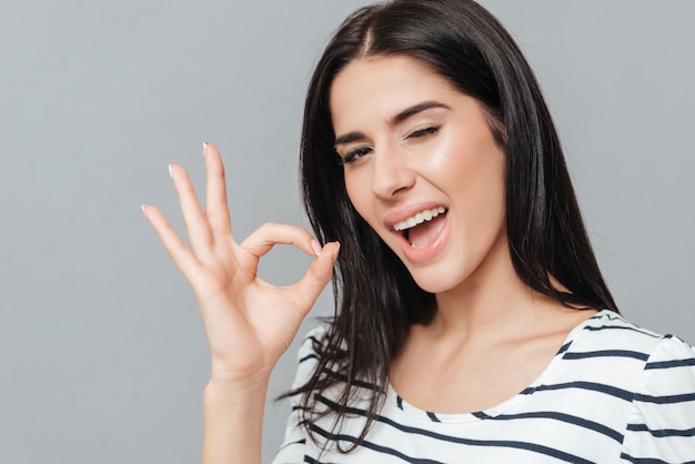 Jeune femme espiègle debout sur une surface grise tout en faisant un geste OK. Regardez devant.