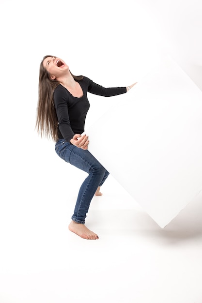 Jeune femme essayant de soulever un cube lourd et criant d'une épreuve. Image isolée sur le mur blanc.