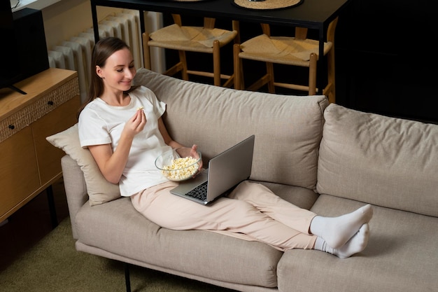 Une jeune femme est allongée sur le canapé et profite du week-end pour regarder des films sur un ordinateur portable et manger du popcor