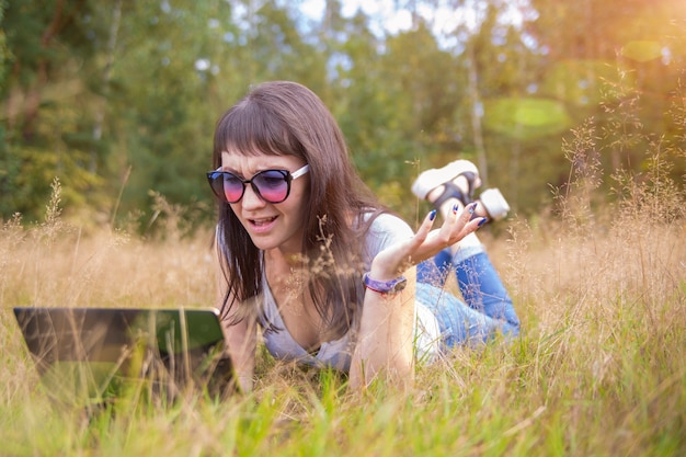 jeune femme est allongée sur l'herbe avec un ordinateur portable et parle avec émotion au téléphone.