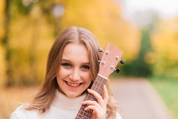 Une jeune femme est assise sur le banc dans le parc et joue du ukulélé, posant avec guitare