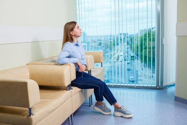 Une jeune femme est assise sur un canapé dans une salle d'attente avec une belle vue sur la ville depuis la fenêtre