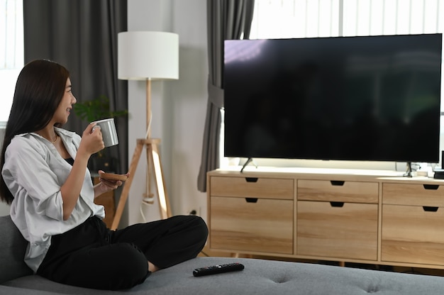 Une jeune femme est assise sur un canapé et regarde la télévision tout en tenant une tasse de café dans les mains