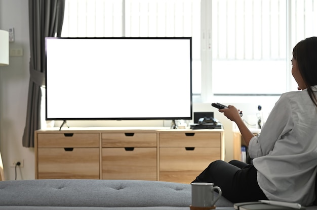 Une jeune femme est assise sur un canapé et tient la télécommande en regardant la télévision à la maison.