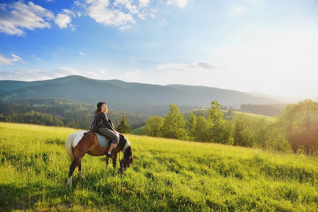 Jeune femme est assise sur un cheval dans les montagnes au coucher du soleil en été