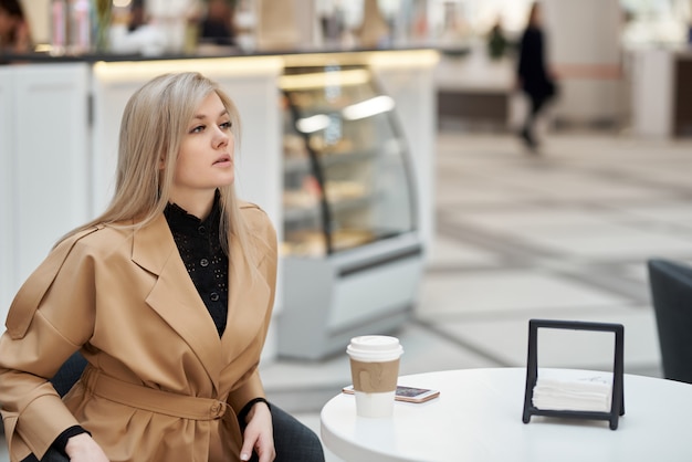 Une jeune femme est assise dans un café, un centre commercial, chat sur téléphone mobile et tenant une tasse de café en papier à la main.