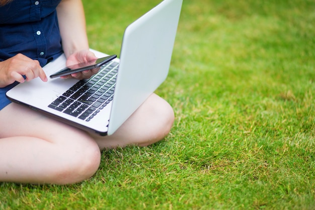 une jeune femme est assise dans un pré avec un ordinateur portable et parle lors d'une conférence en ligne au travail