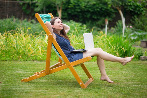 une jeune femme est assise dans un pré avec un ordinateur portable et parle lors d'une conférence en ligne au travail