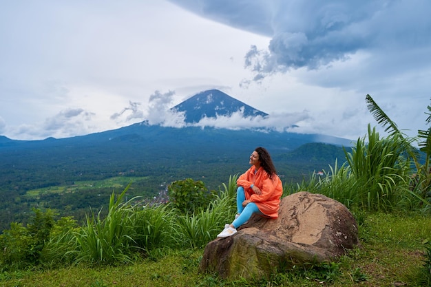 Une jeune femme est assise sur un grand rocher dans un point de vue et profite de la vue du volcan sacré du mont Agung caché par les nuages un jour de pluie sur l'île de Bali