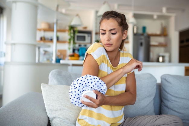 Une jeune femme est assise à la maison sur son canapé et touche son coude tout en souffrant de douleurs au coude en plaçant un paquet de glace sur le point blessé