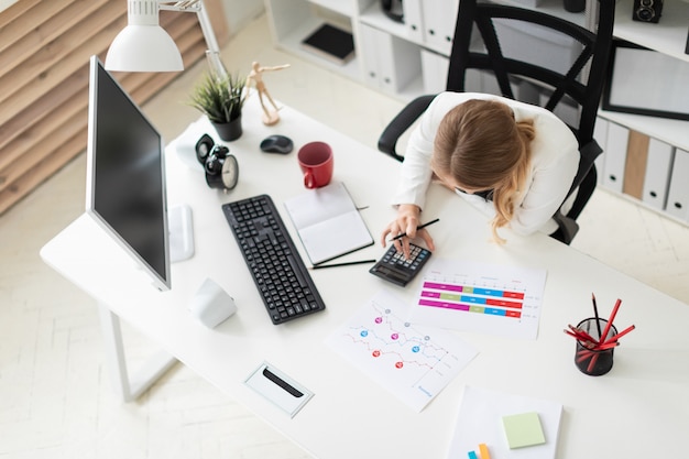 Une jeune femme est assise à un ordinateur dans le bureau et compte sur une calculatrice.