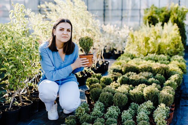 Une jeune femme est assise près d'un parterre de fleurs dans la rue près d'un magasin de plantes et choisit un pot avec un petit arbre Une femme choisit des plantes pour aménager la cour Concept de jardinage domestique